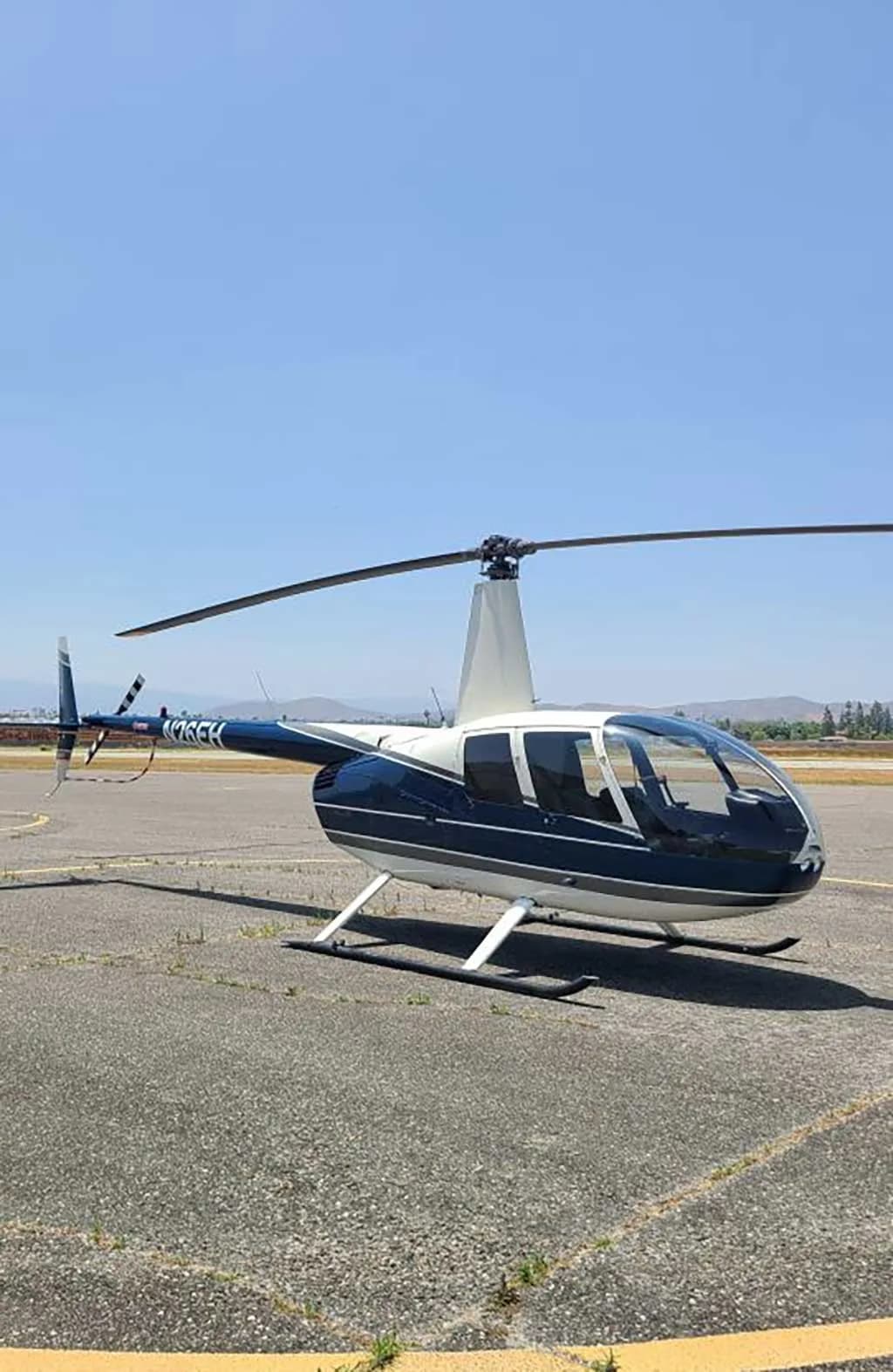 A blue and white Robinson R44 helicopter, registration N26EH, is parked on an asphalt tarmac under a clear blue sky.