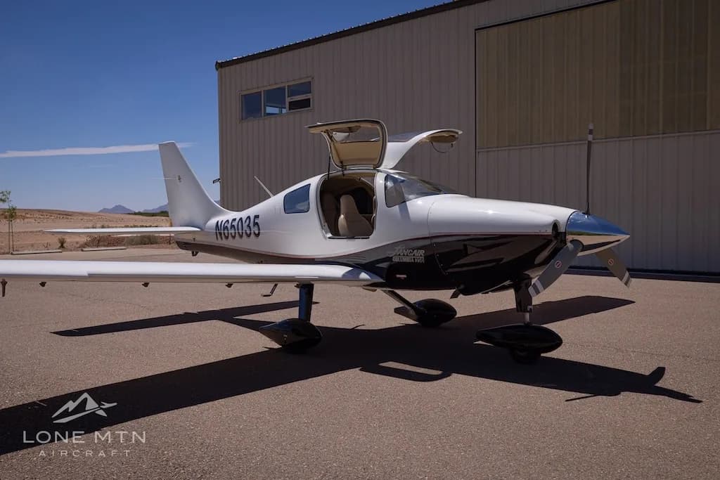 A white and black single-engine aircraft, N65035, with its gull-wing doors open, parked on an airport ramp with a hangar in the background.