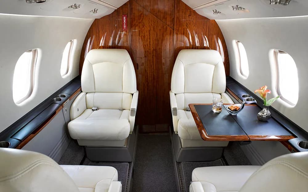 The private jet cabin interior featuring cream leather seats, a wooden bulkhead, and a table with a drink and flowers.