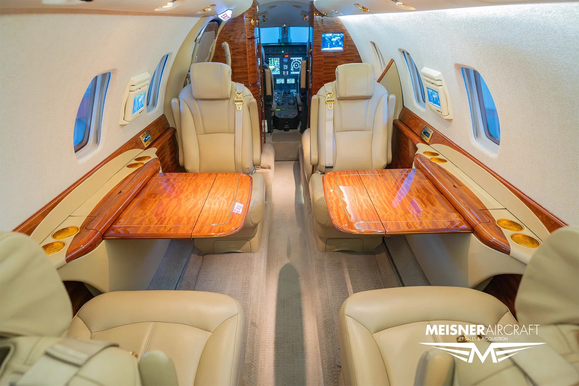 The interior cabin of a business jet showing beige leather seats with wooden tables deployed between them.