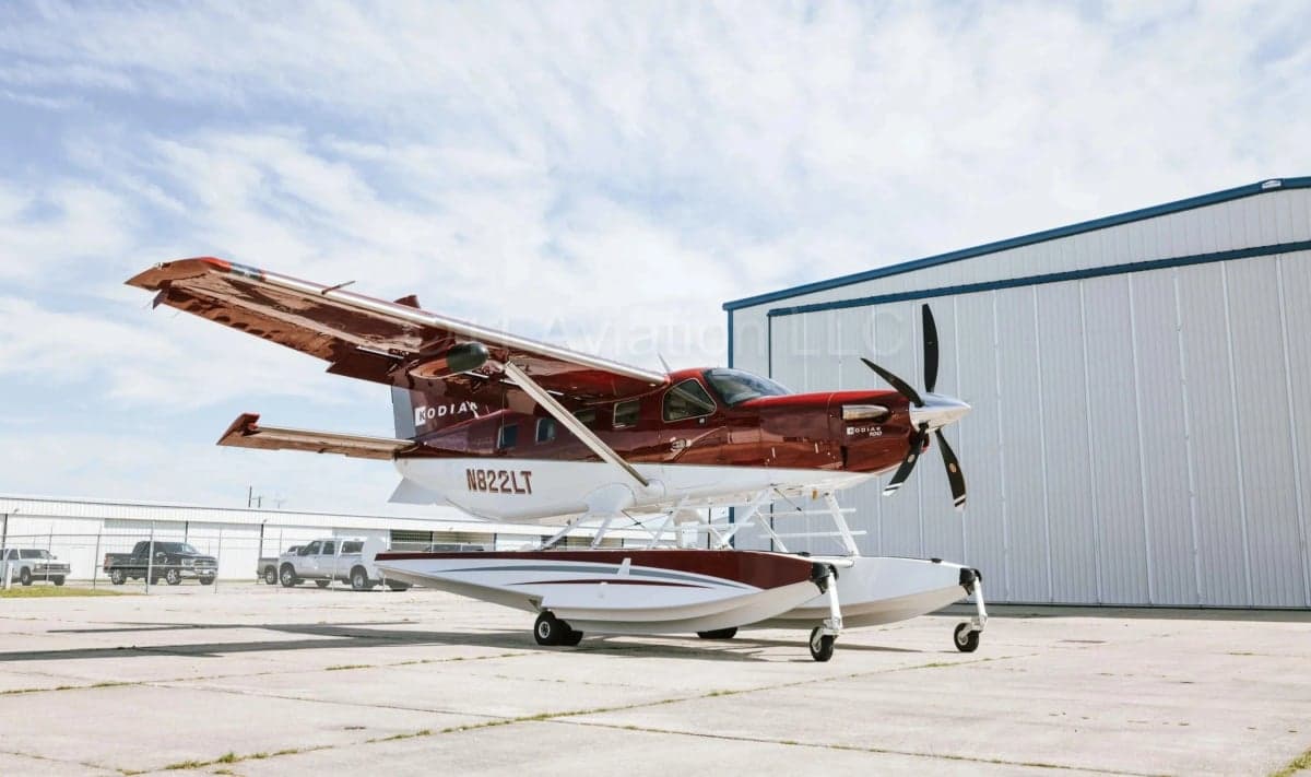 A full side view of a red and white Kodiak 100 seaplane with floats, parked on an airfield next to a hangar.