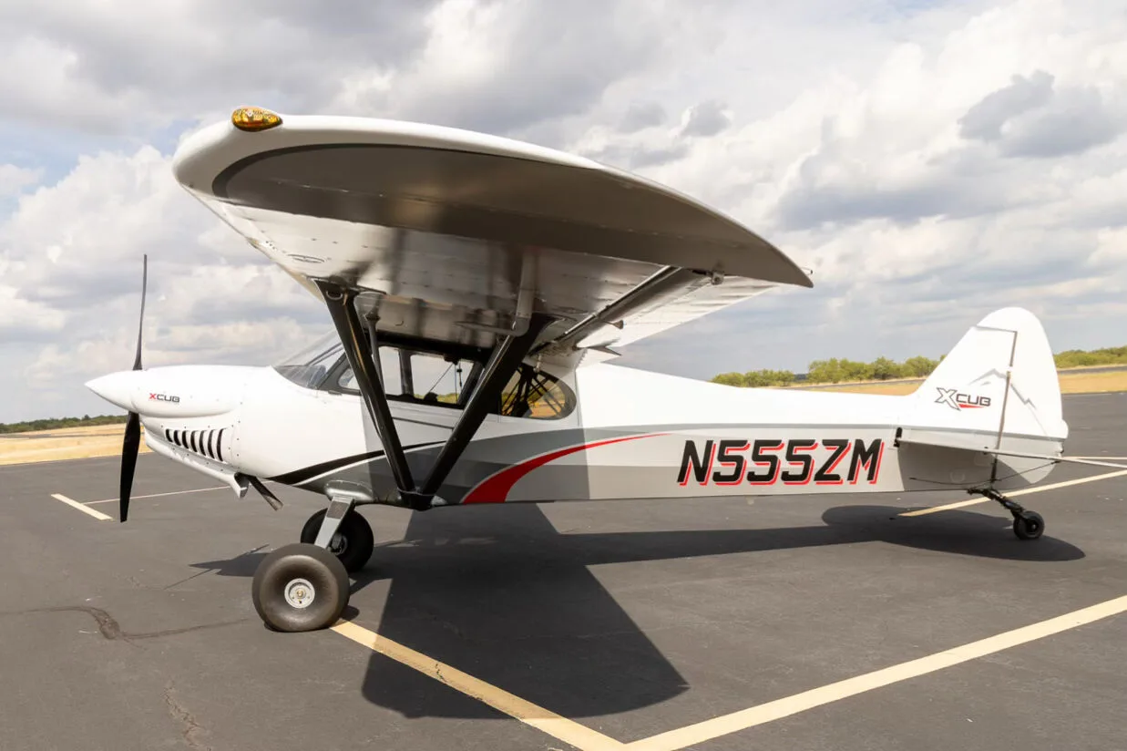 A side view of the white and grey X-Cub aircraft N555ZM with red accents, parked on an asphalt runway.