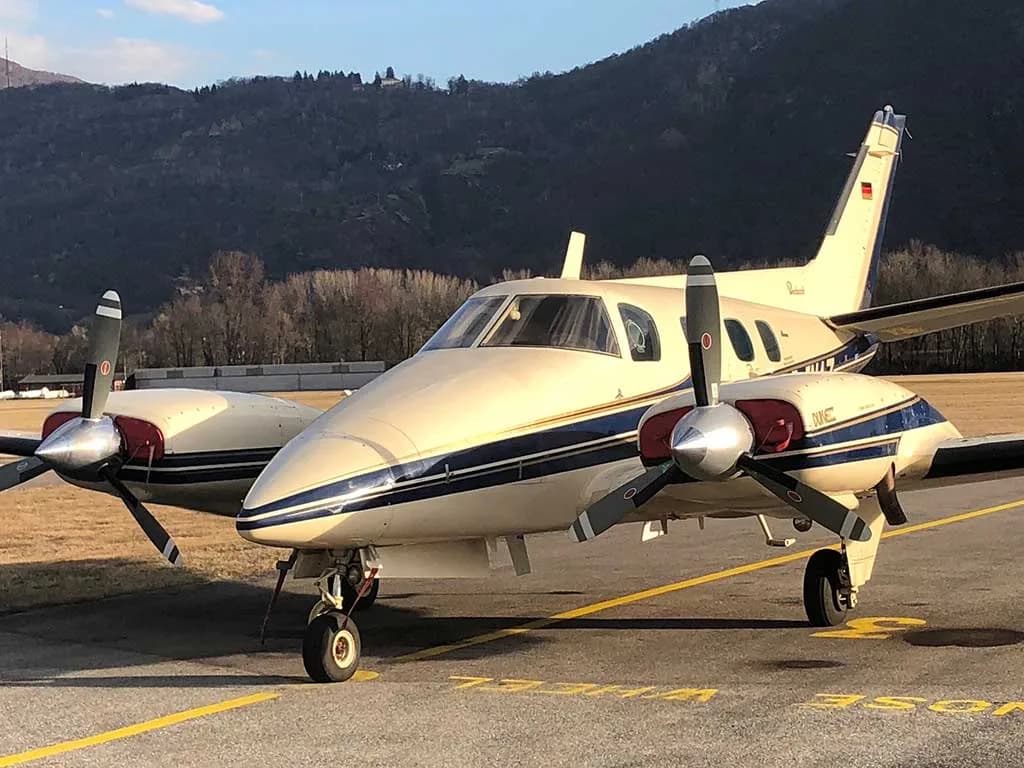A beige and blue twin-engine Piper PA-31-350 Navajo Chieftain aircraft is parked on an asphalt runway with mountains in the background.