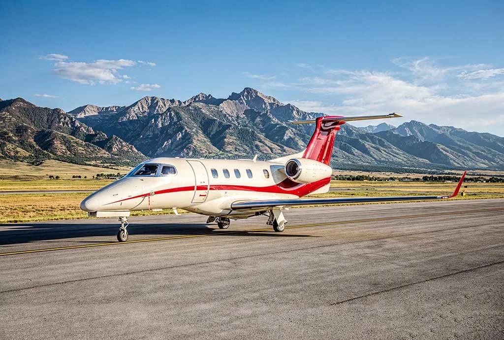 A white and red Embraer Phenom 300 private jet on a runway with mountains in the background.