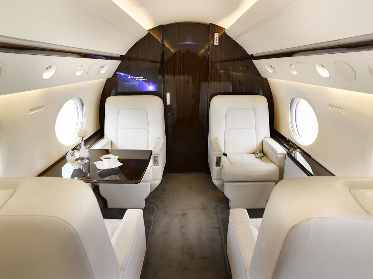 The luxurious interior of a Gulfstream G650 cabin, featuring white leather seats, a dark wood table, and a monitor.