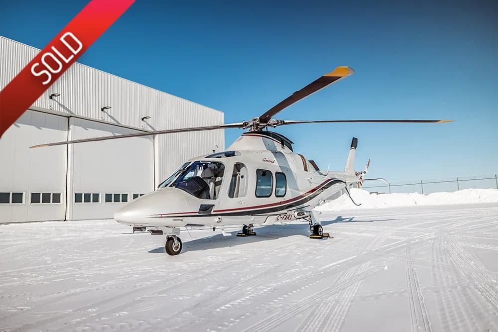 A white AgustaWestland AW109 Grand helicopter with red and black stripes sits on a snowy tarmac in front of a hangar.