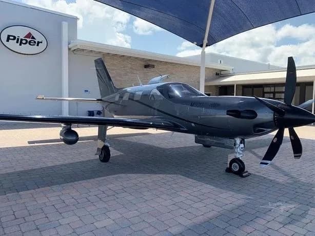 A dark grey Piper M600/SLS turboprop aircraft parked on a paved area in front of a building with a "Piper" logo.