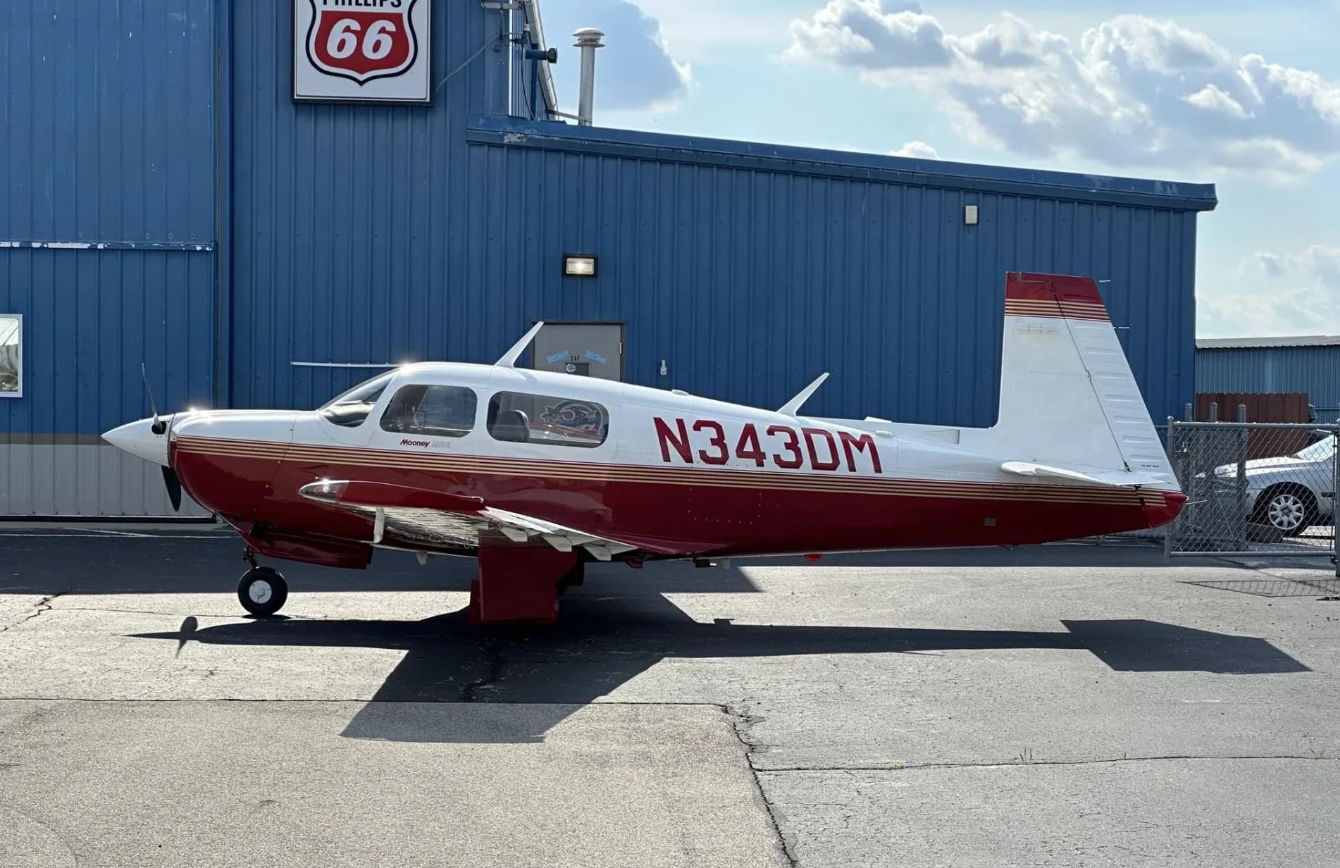 A full side view of a red and white Mooney aircraft (N343DM) parked in front of a blue hangar with a Phillips 66 sign.