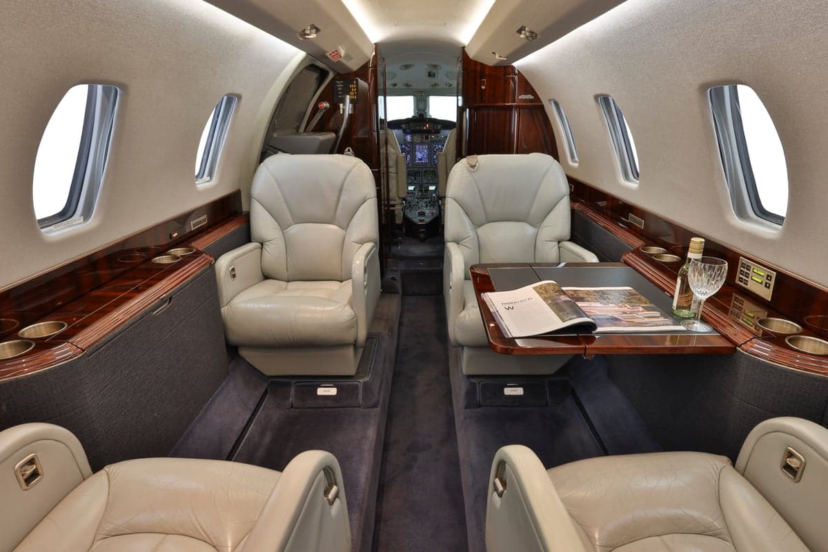 The interior of a Citation CJ3 aircraft, showing the cabin seats, a table with a magazine and wine bottle, and the cockpit in the background.