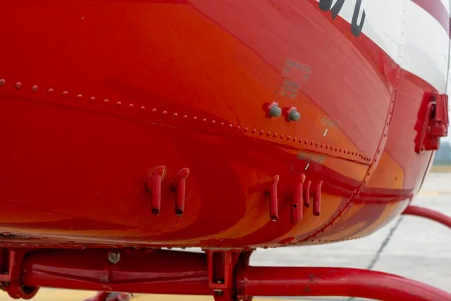 A close-up view of the red fuselage of a helicopter, showing rivets and small external components.