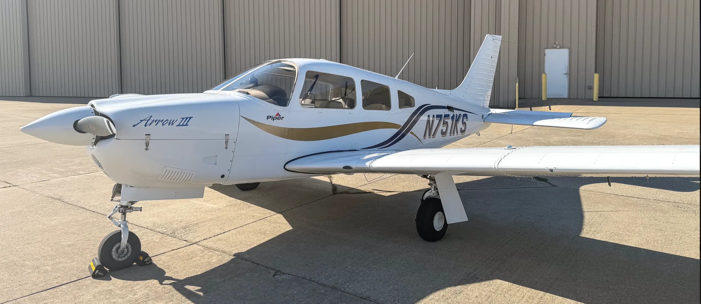 A front three-quarter view of the white Piper Arrow III aircraft, tail number N751KS, parked on an airport tarmac in front of a hangar.