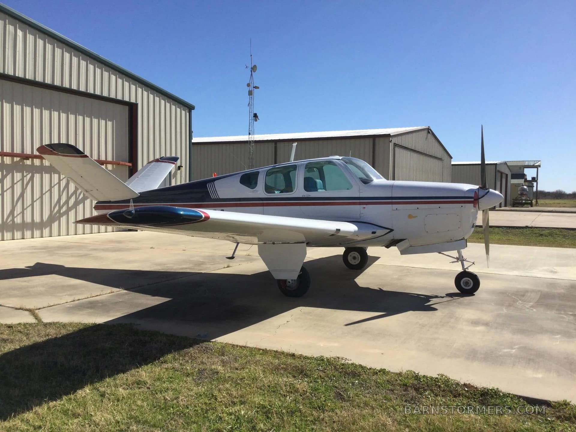 A white aircraft with blue and red stripes is parked on a concrete pad with grass nearby, next to hangars under a clear blue sky.