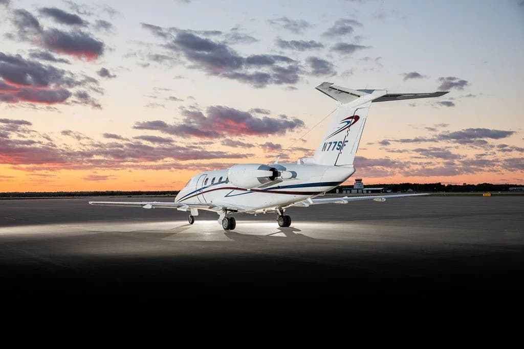 A white Cessna Citation business jet with blue and red stripes on the tail, viewed from the rear-right on a tarmac at sunset.
