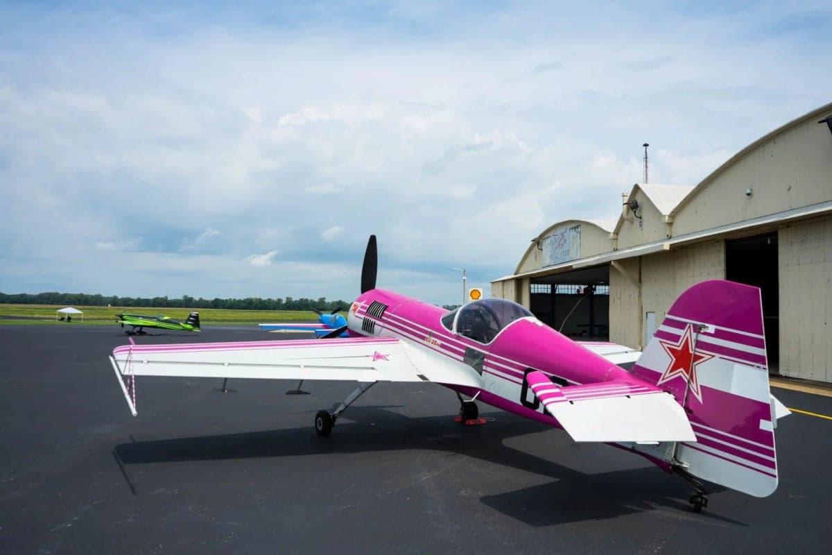 Rear quarter view of a pink and white Sukhoi Su-26 aircraft parked on the tarmac near a hangar.