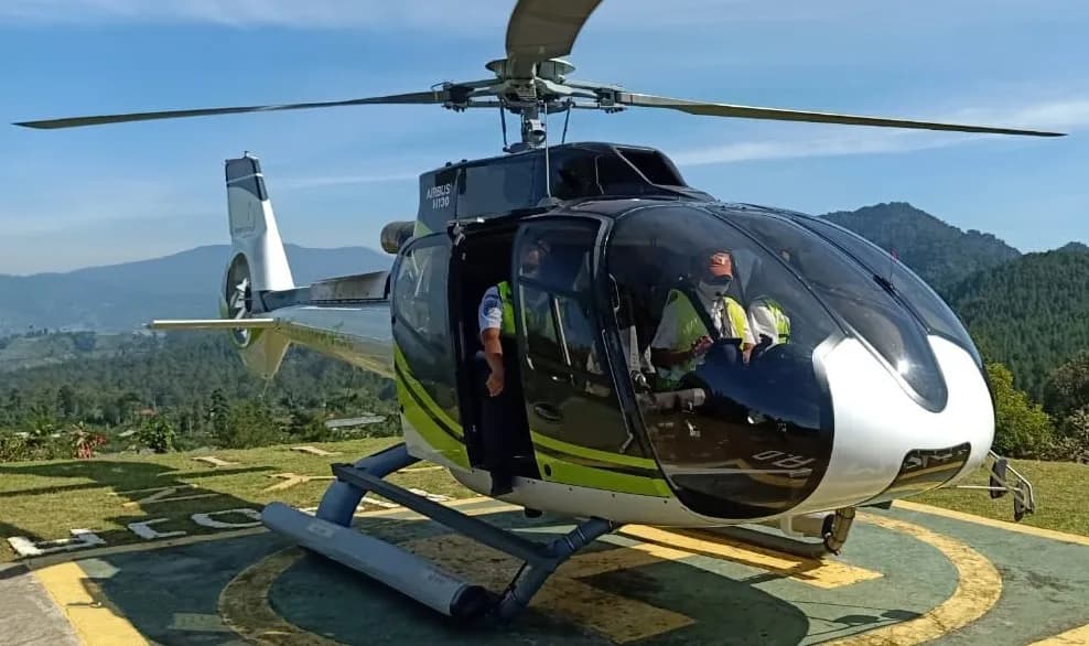 An Airbus H130 helicopter on a helipad with mountains in the background.