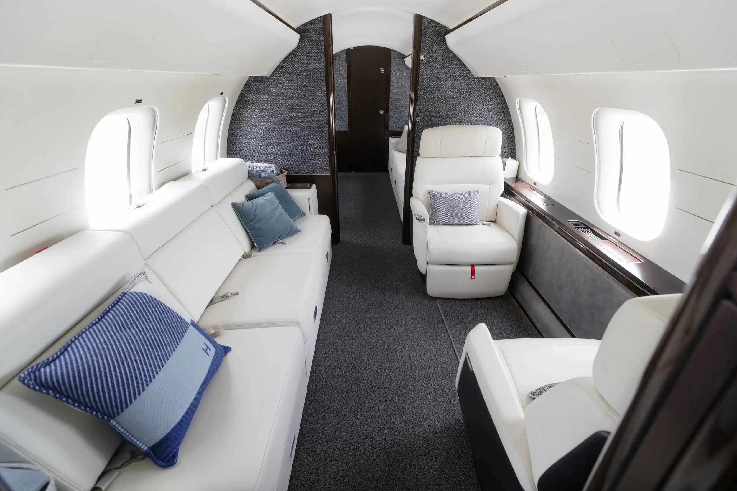 The spacious aircraft cabin features white leather seating, including a long sofa and individual chairs, with blue accent pillows.