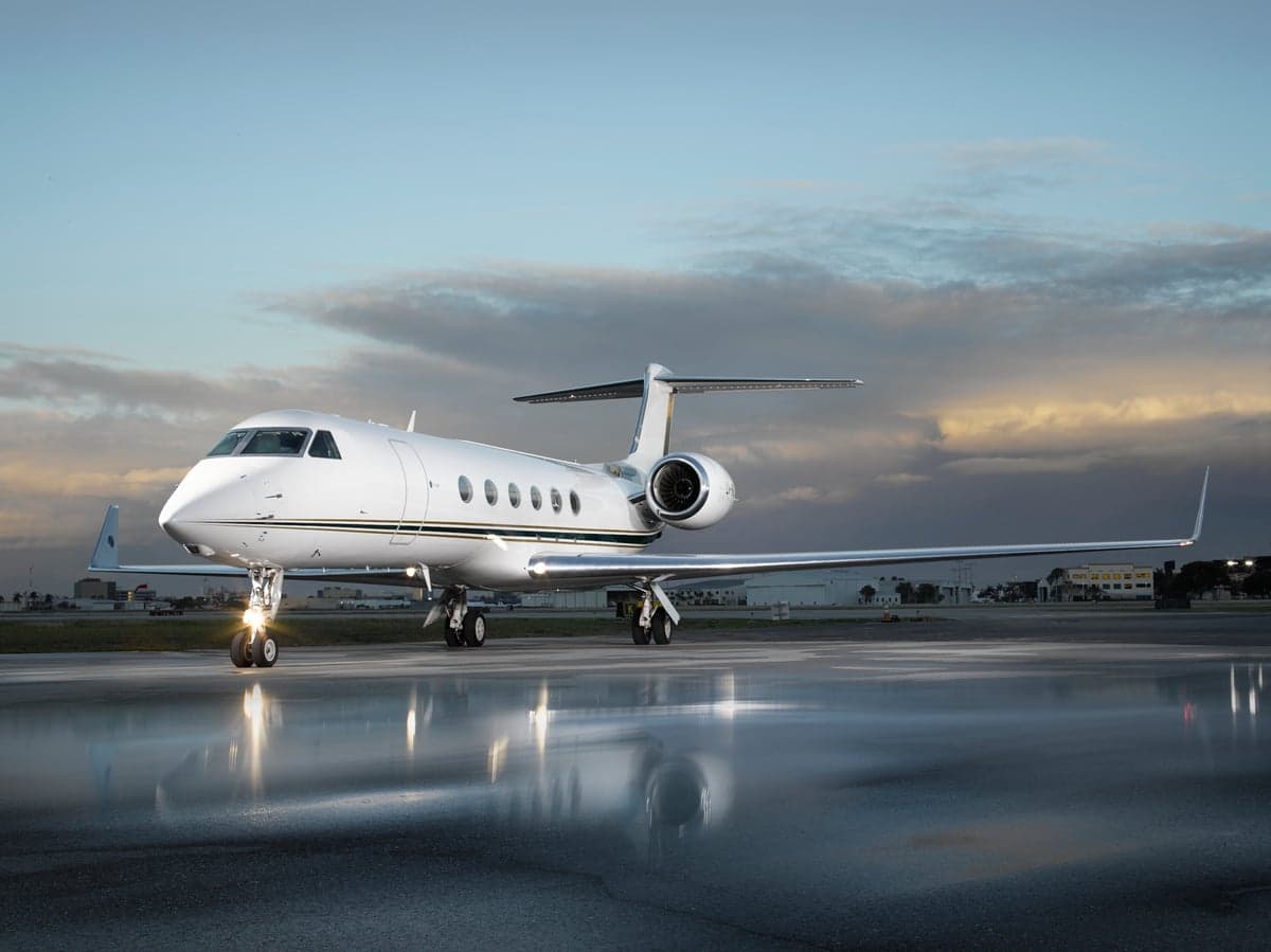 Gulfstream G550 aircraft with landing lights on parked on a wet tarmac reflecting the sky.