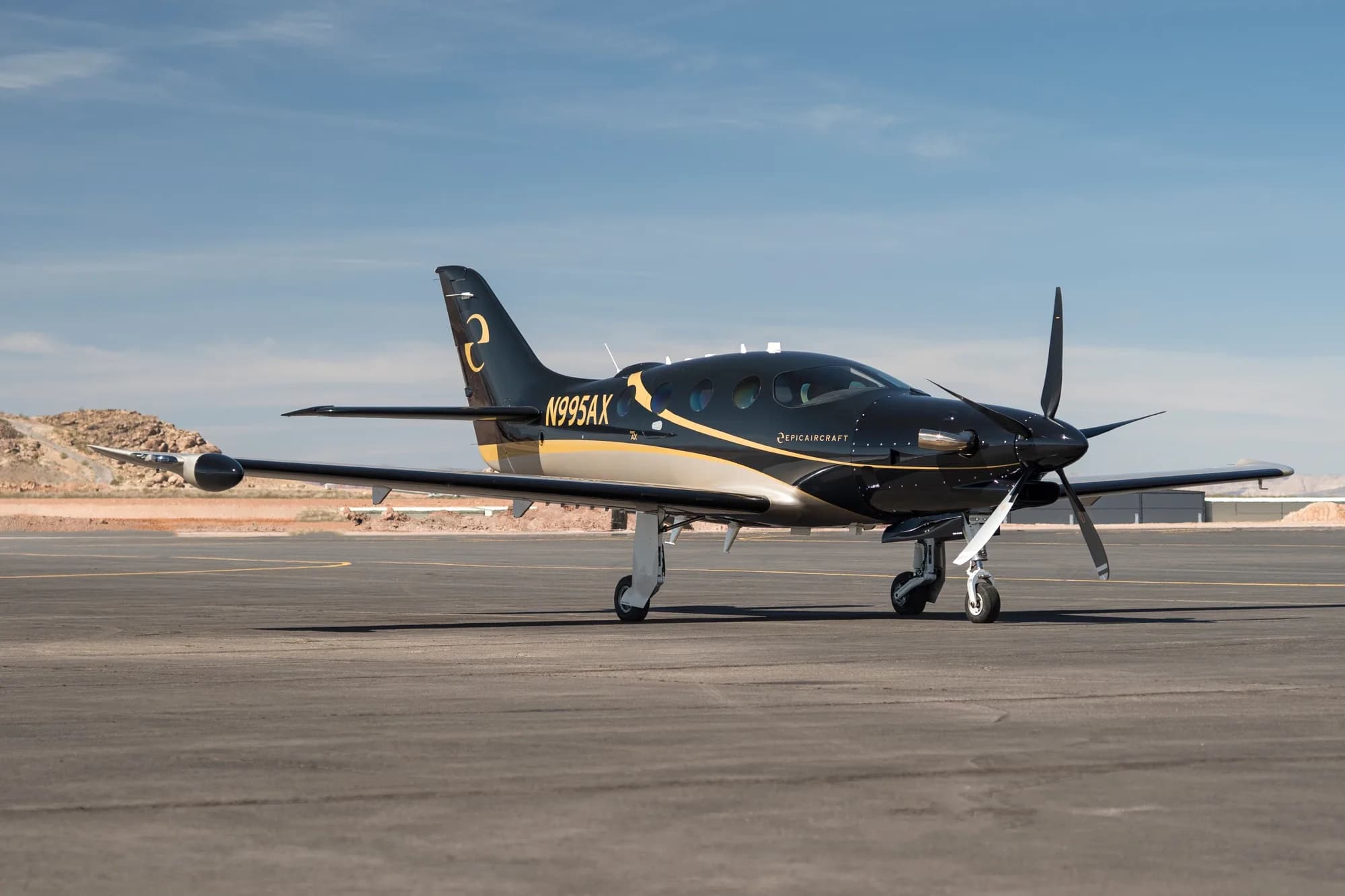 A black and gold Epic E1000 GX aircraft (N995AX) is parked on a tarmac with rocky terrain in the background.