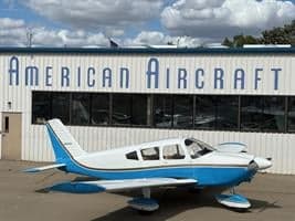 A white and blue Piper PA-28 aircraft is parked on a tarmac in front of the American Aircraft building.