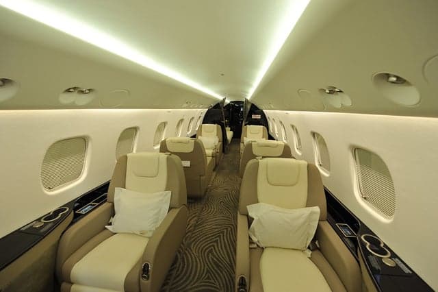 The luxurious interior cabin of a private jet with beige and cream leather seats and overhead lighting.