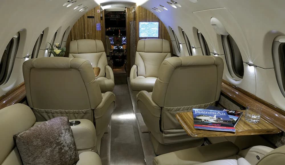 The spacious interior cabin of a business jet featuring beige leather seats and a magazine on a wooden table.