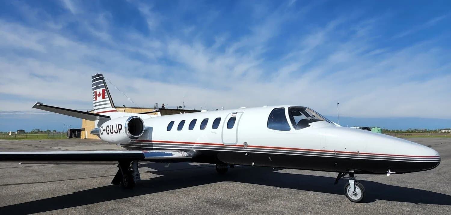 A white and black Cessna Citation business jet with red and grey stripes and a Canadian flag on the tail, parked on an airport apron under a blue sky.