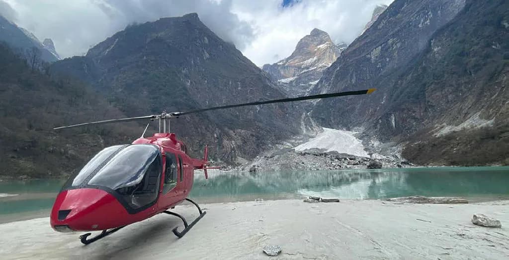 A red helicopter is parked on a light-colored shore next to a glacial lake, with a large mountain range in the background.