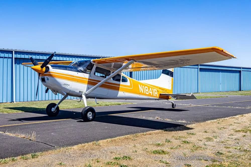 A white and orange single-engine propeller aircraft with tail number N1841R parked on an asphalt ramp in front of blue hangars.