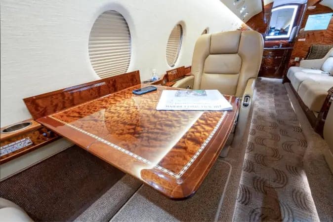A close-up view of a light-colored leather seat and a polished wood table with a newspaper on it inside a private jet cabin.