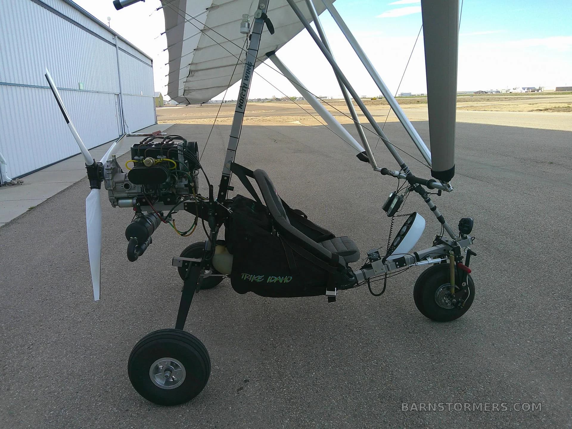 A black and white ultralight aircraft with a single propeller is parked on an asphalt runway next to a white hangar.
