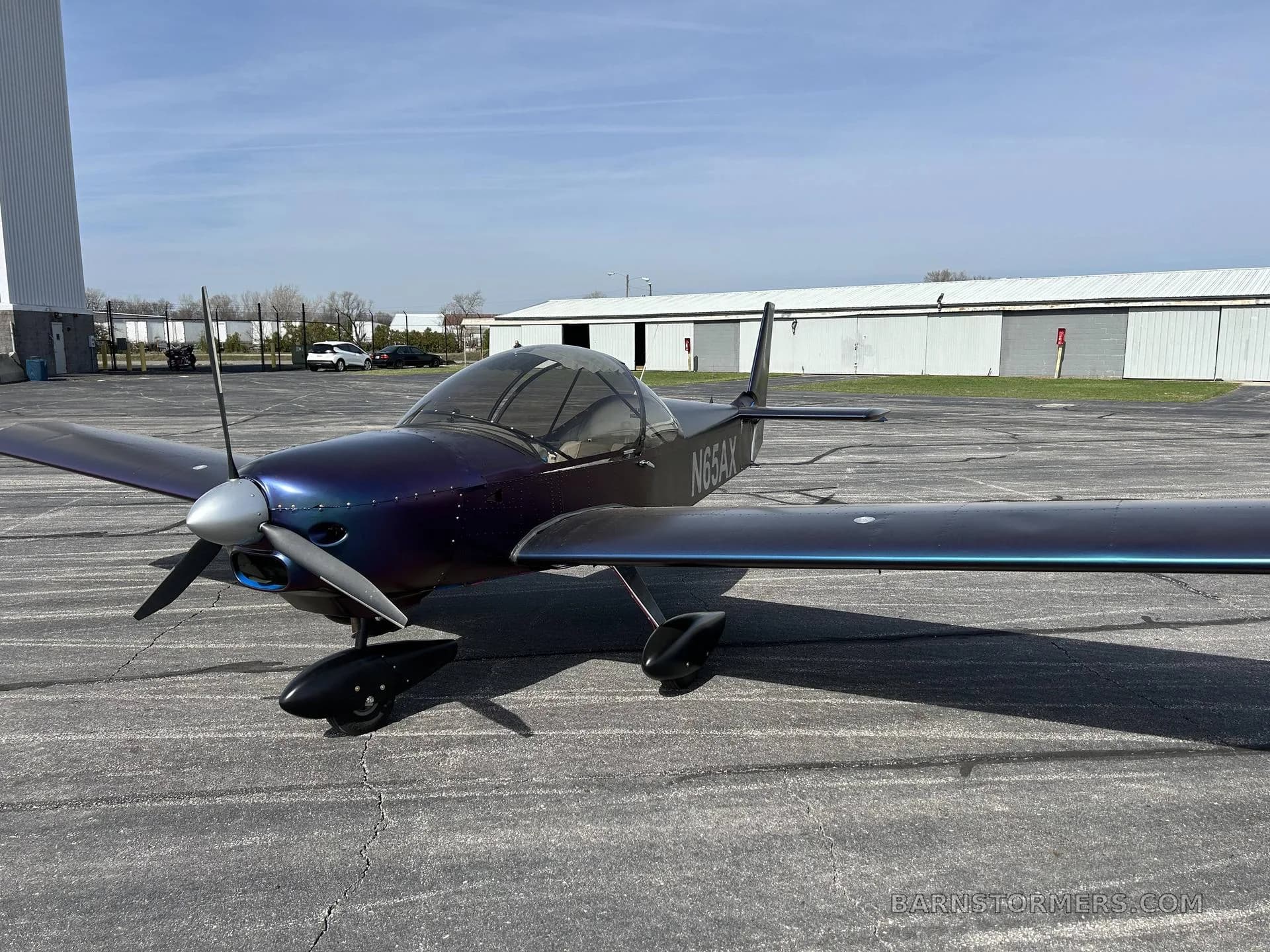 A front-left view of a purple and blue iridescent Zenith aircraft (N65AX) parked on an airport tarmac with hangars in the background.
