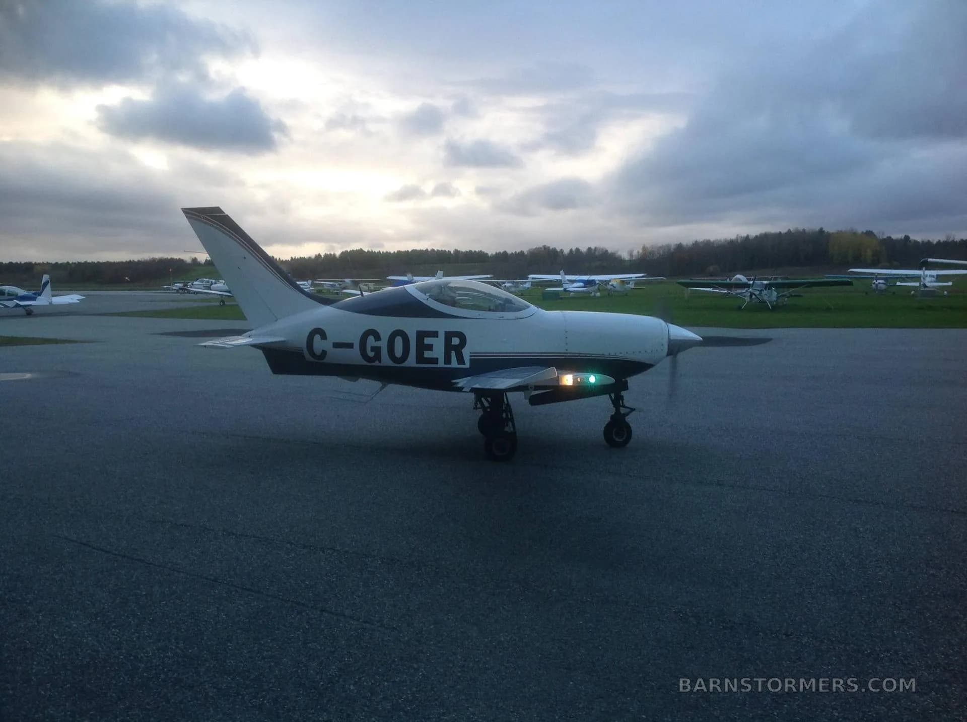 A white and blue single-engine aircraft with its landing lights on, taxiing on an airfield under a cloudy sky, from a slightly different angle.