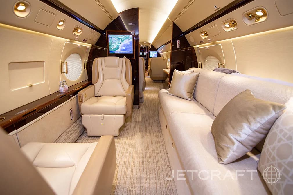 A private jet interior with a beige sofa, a single seat, and a large screen displaying a map.