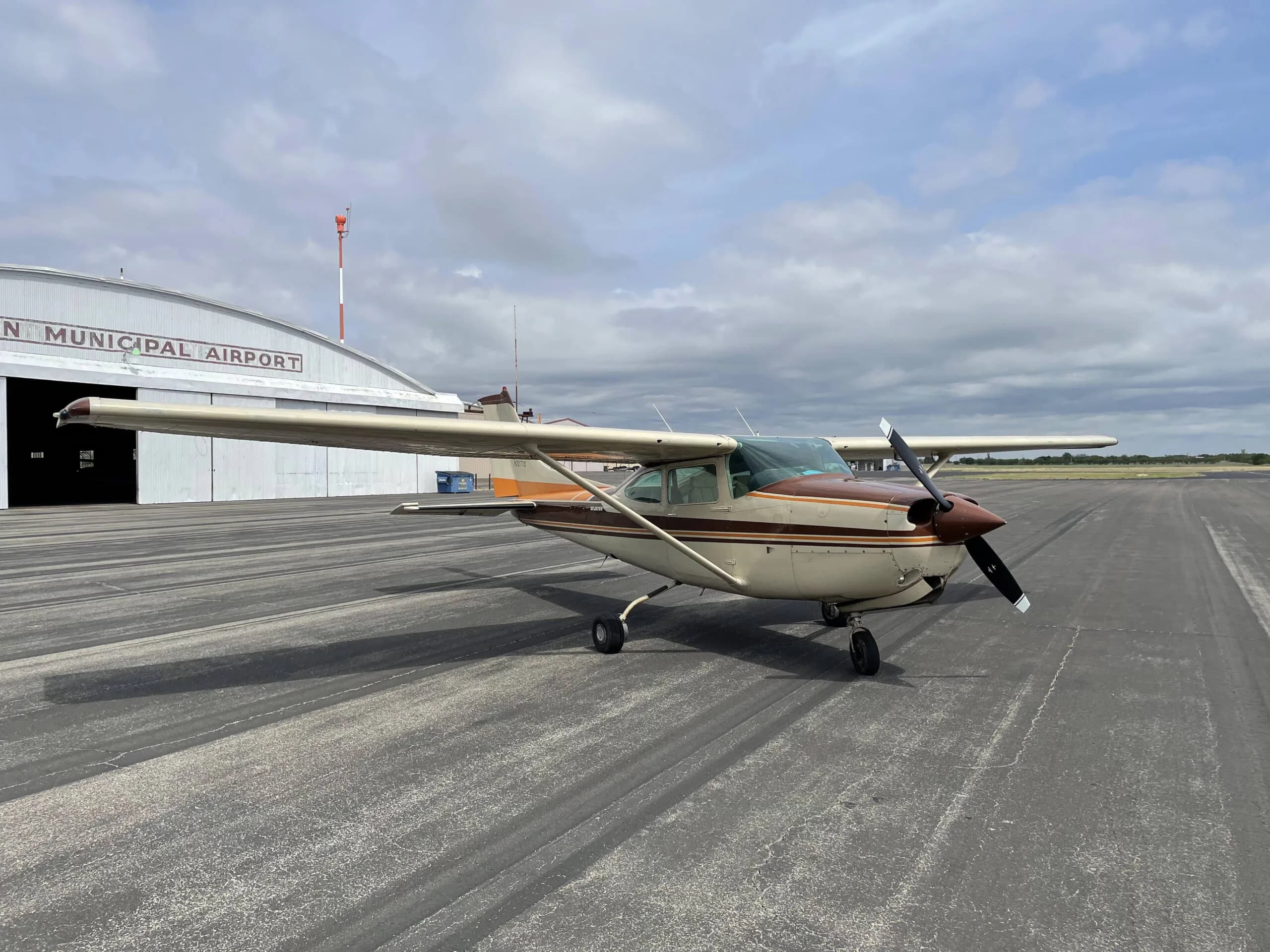 A cream and brown single-engine propeller aircraft, likely a Cessna, parked on an airport tarmac with a hangar in the background.