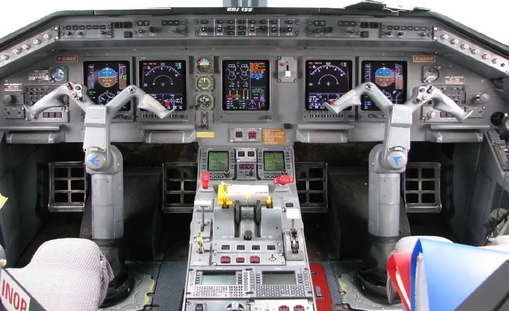The cockpit of an ERJ 135 aircraft, showing the instrument panel with multiple digital displays, yokes, and the central throttle quadrant.