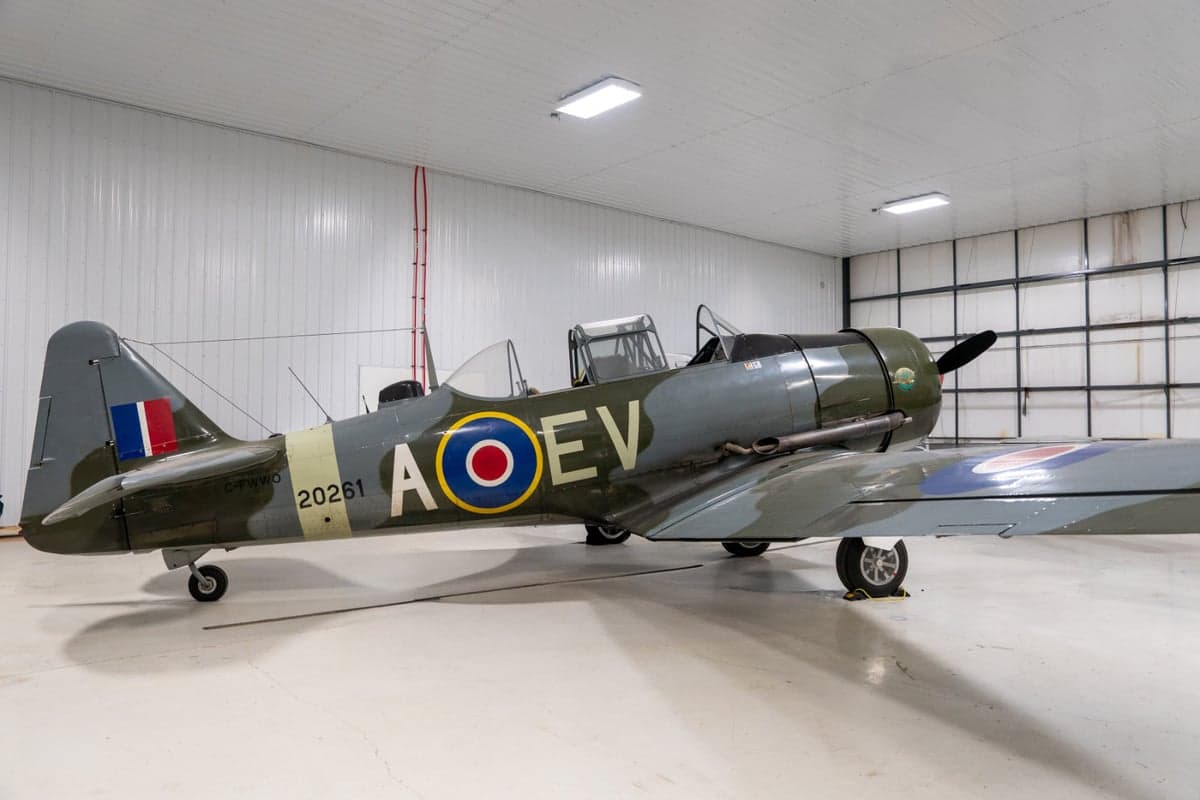 A full side view of a vintage military trainer aircraft with a camouflage paint scheme and open canopies, parked in a white hangar.