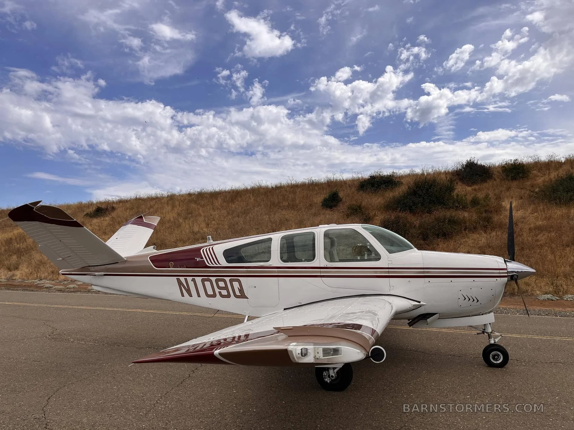 A white and brown Beechcraft Bonanza with registration N109Q, parked on an asphalt surface with a dry grassy hill under a blue sky.