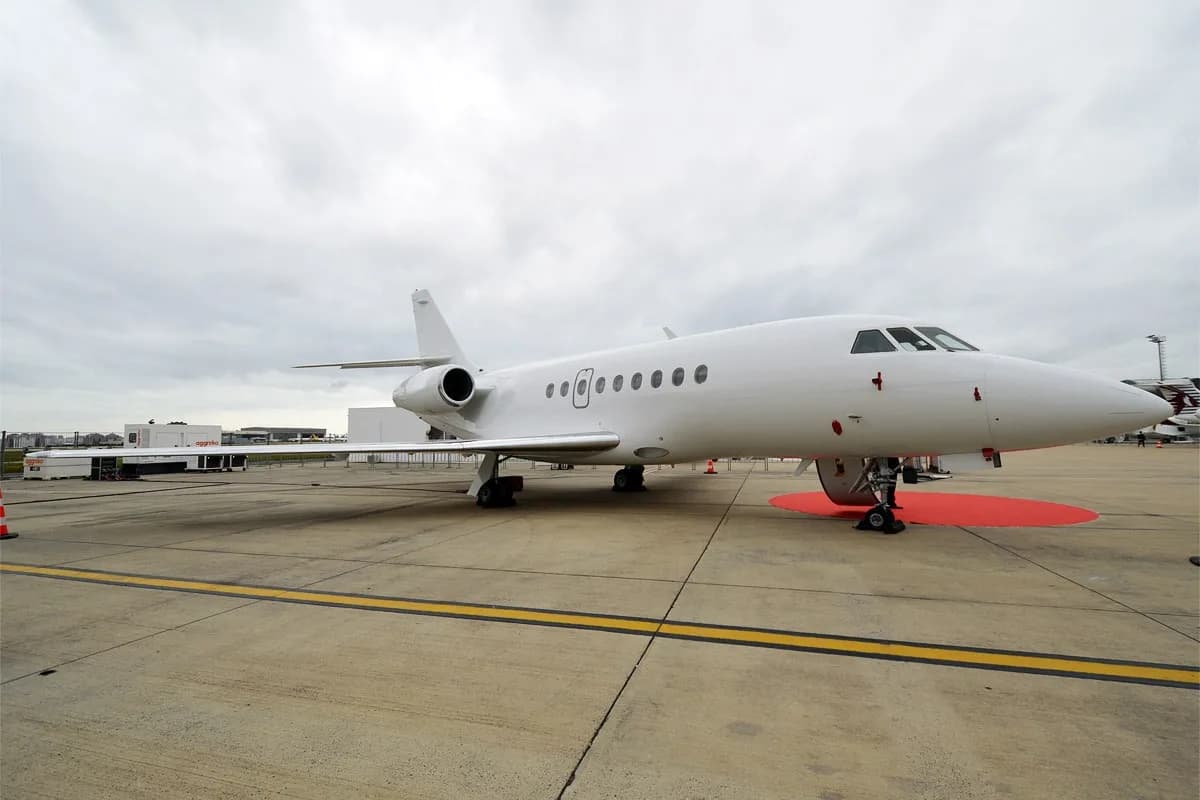 A white Dassault Falcon private jet parked on an airport tarmac under an overcast sky.