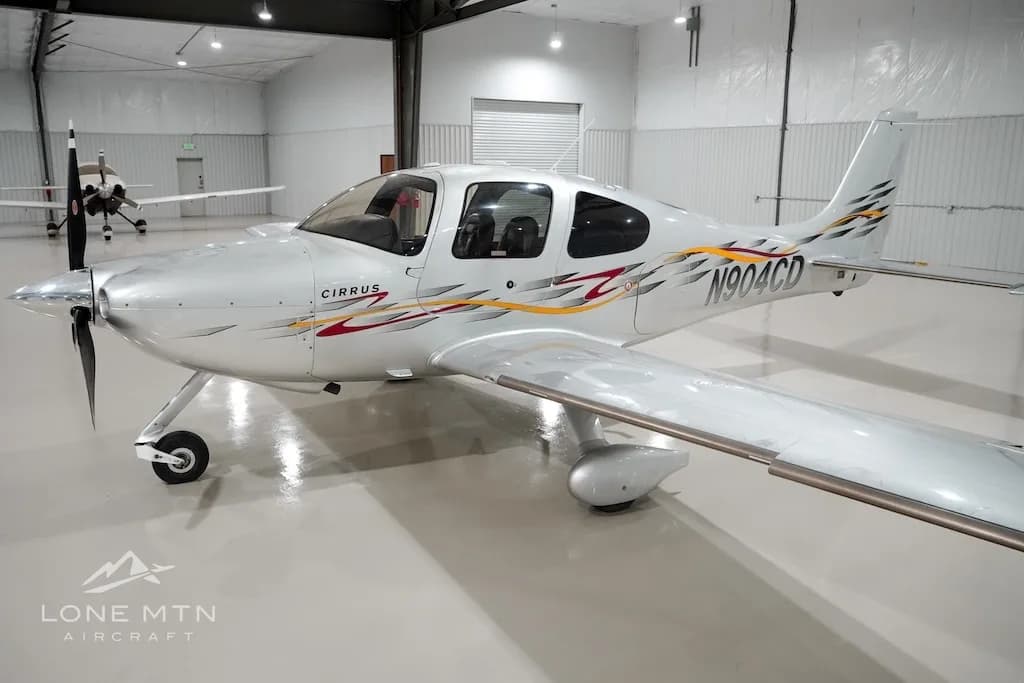 A silver Cirrus aircraft with red and yellow accent stripes, registration N904CD, parked in a hangar.