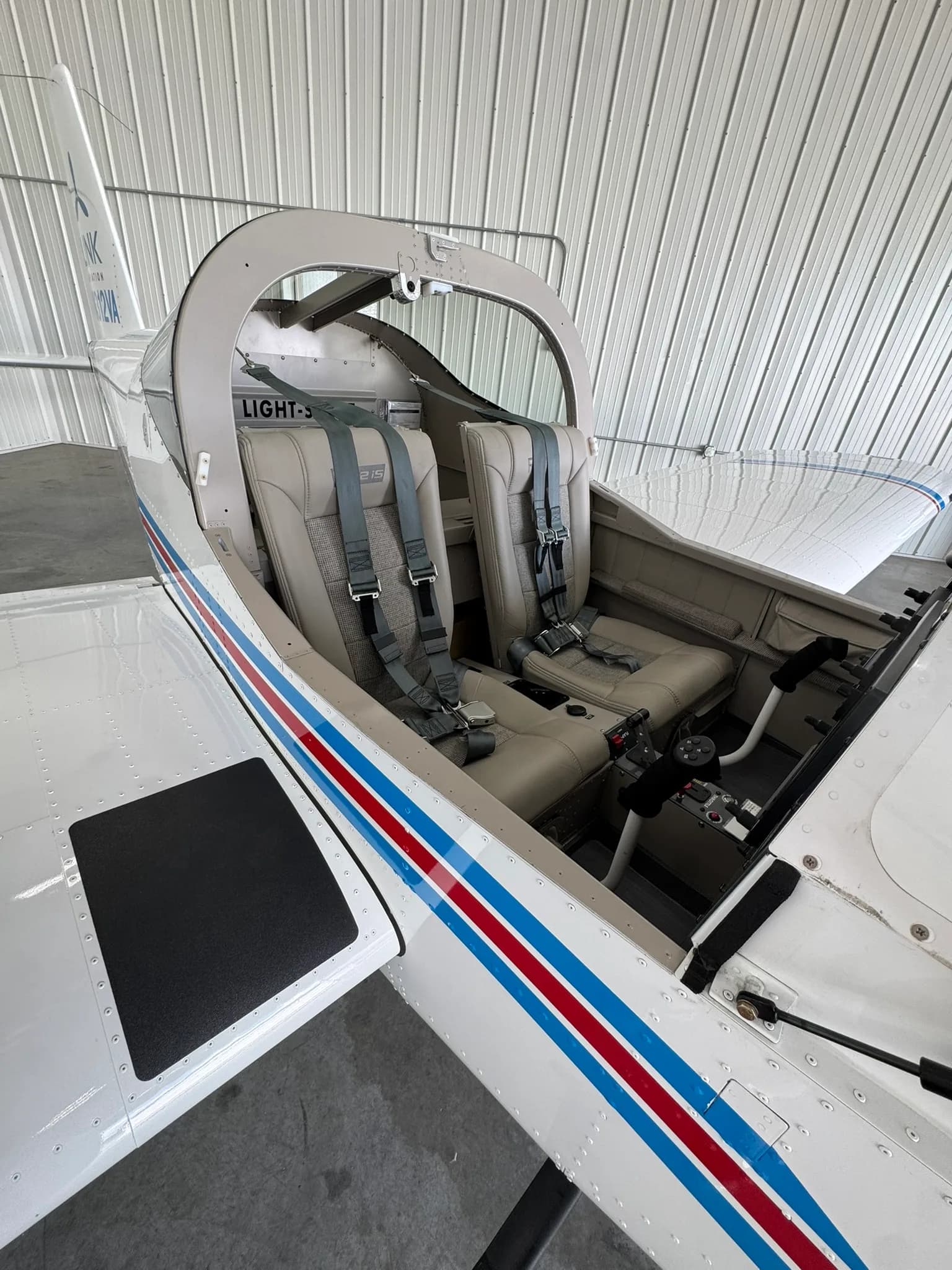 The two-seat interior of a light-sport aircraft, showing beige seats with grey fabric inserts and seatbelts.