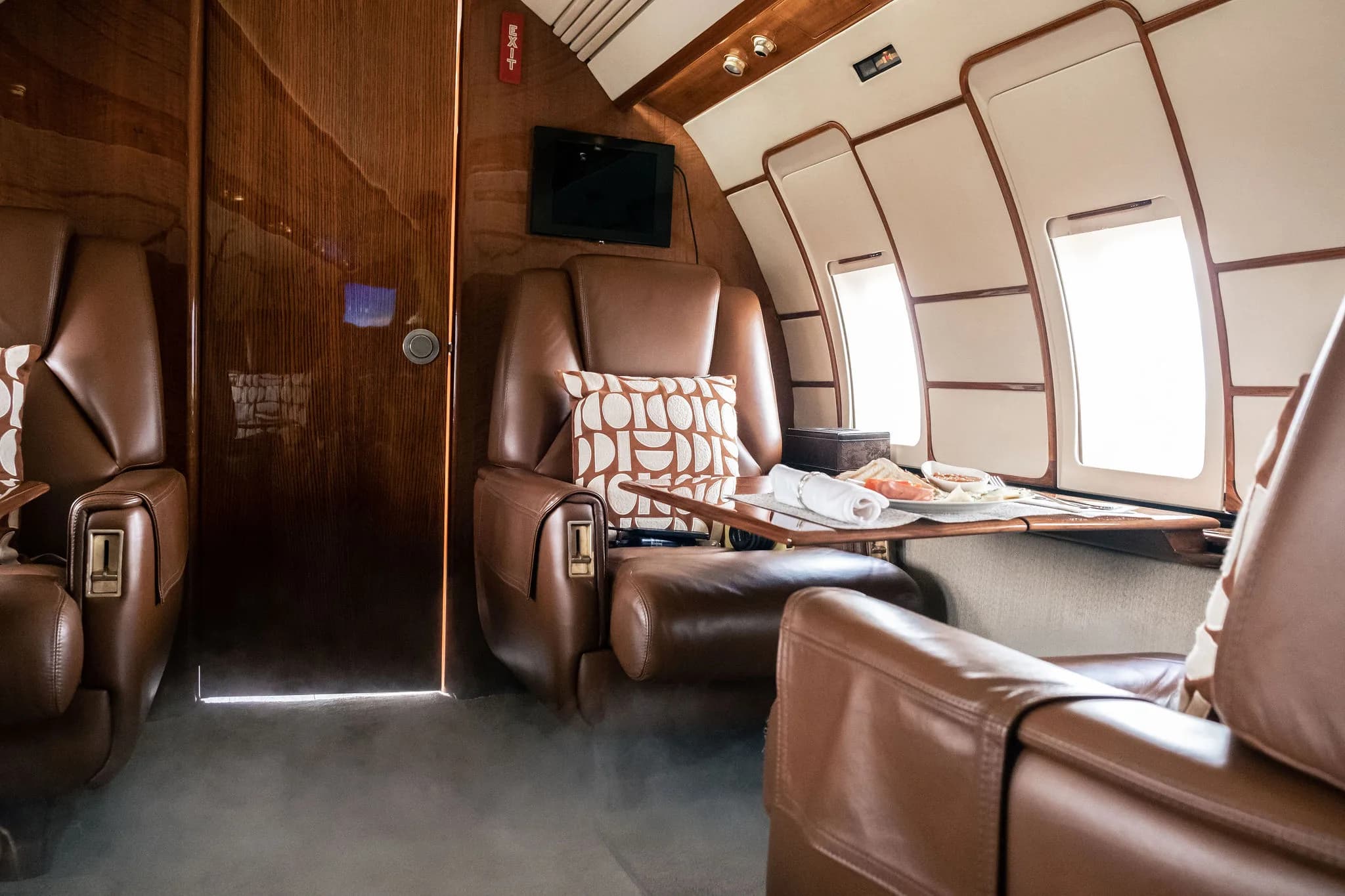 Private jet cabin with brown leather seats, a table set with snacks, and a wooden partition.