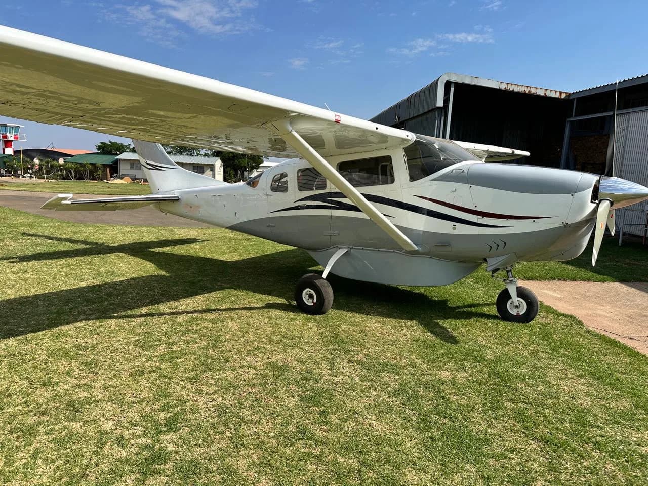 A white and grey single-engine high-wing aircraft parked on green grass next to a hangar.