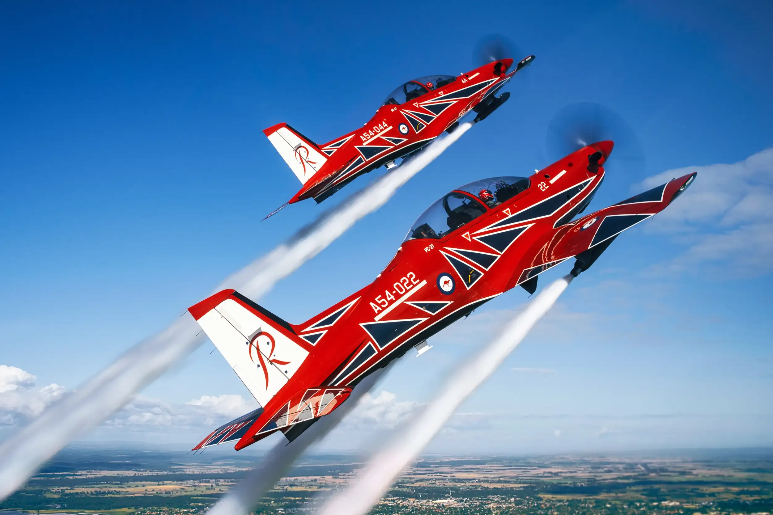 Two red and white Pilatus PC-21 aircraft flying in formation, leaving smoke trails.