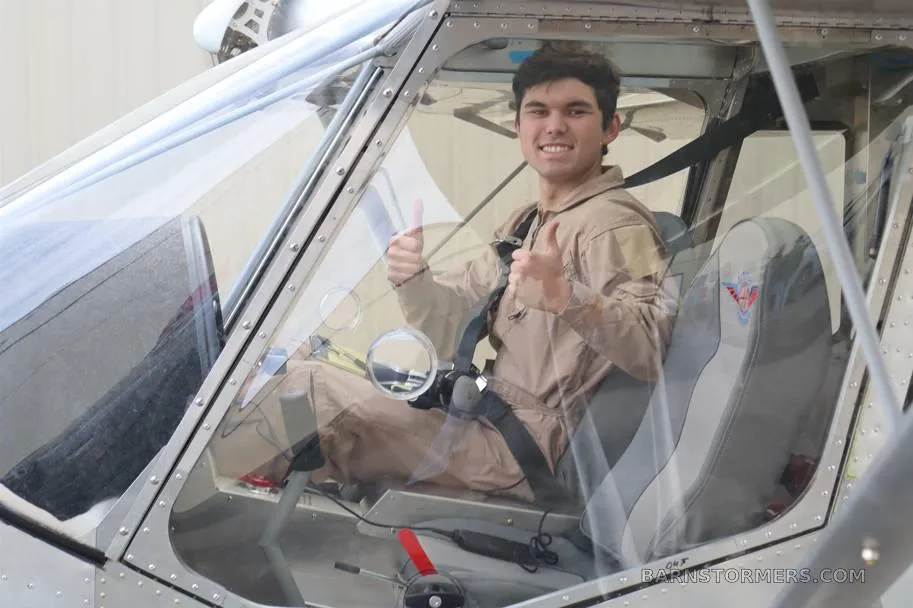 A young man in a flight suit gives two thumbs up from the clear canopy of a light aircraft.