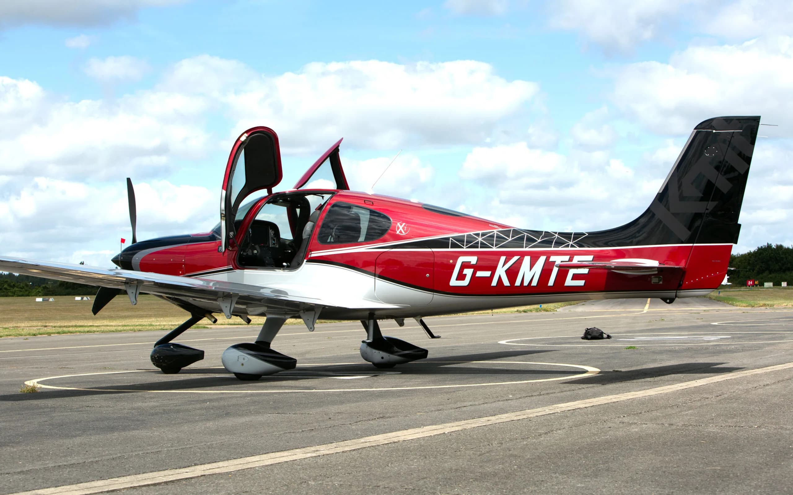 A side view of a red, black, and silver Cirrus SR22 aircraft with registration G-KMTE and open doors, parked on an airfield.