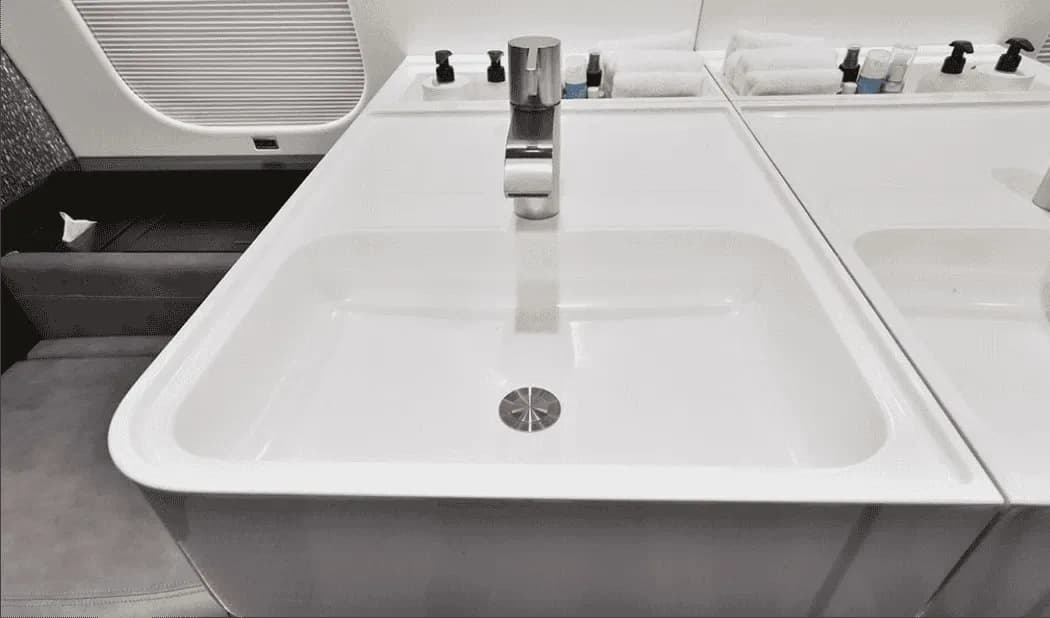 A close-up view of a sleek white sink and faucet in an aircraft lavatory, with toiletries on the counter.
