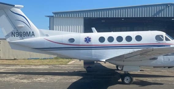 A white twin-turboprop aircraft with red and blue stripes and a Star of Life symbol, parked on an tarmac next to a hangar.