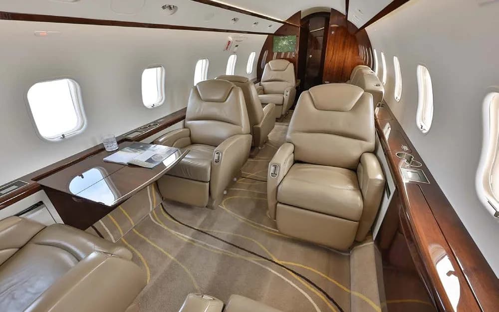 An angled view of the private jet's cabin, featuring tan leather seating, a table, and large windows.