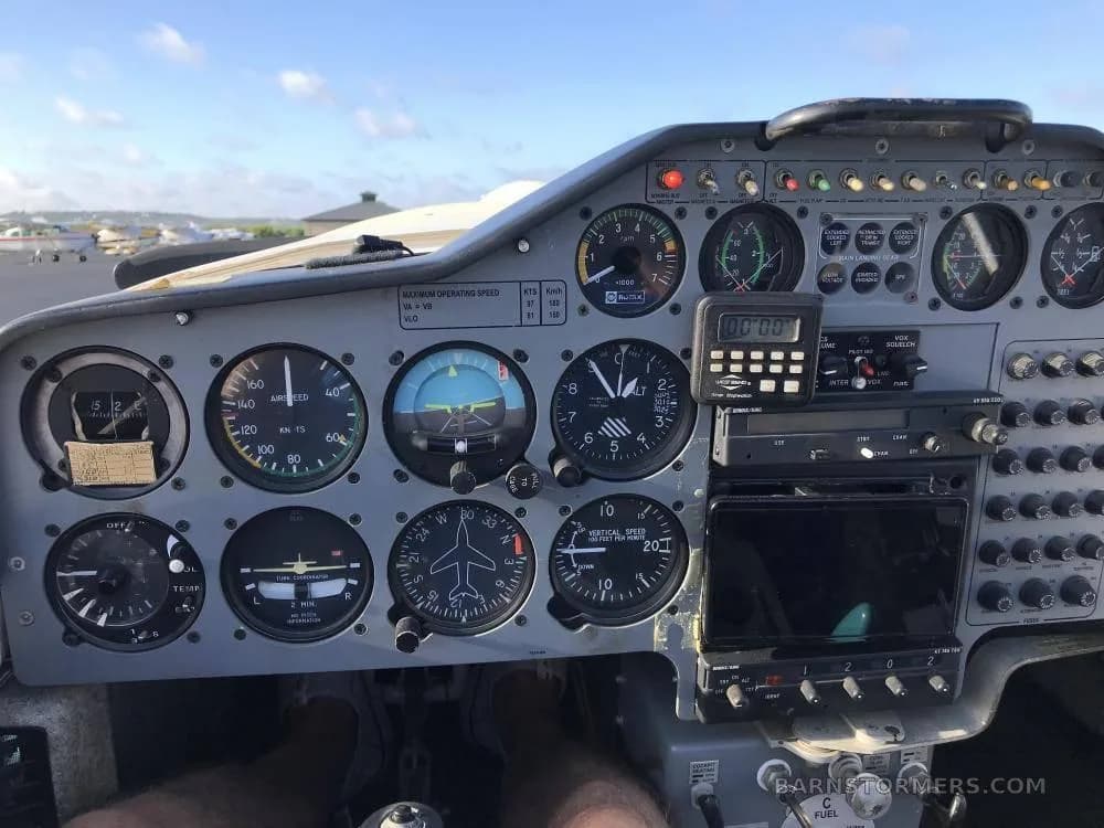The instrument panel of a motorglider features analog gauges, a digital display, and various switches, with a view of the tarmac outside.