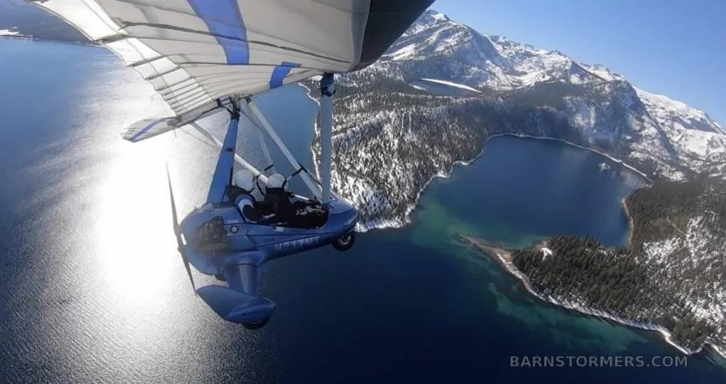 A blue ultralight aircraft with a white and blue wing flies over a large lake surrounded by snow-capped mountains.